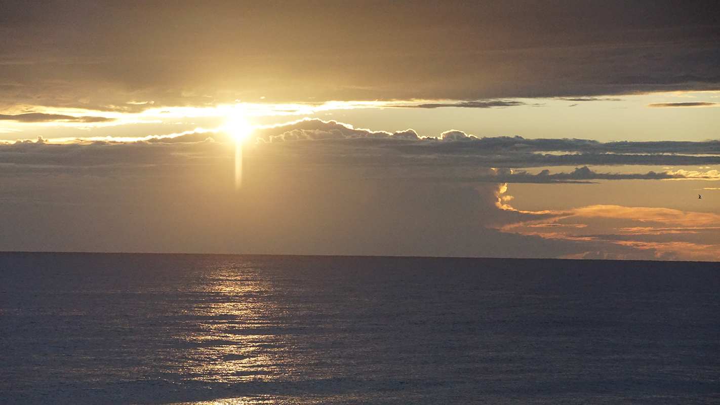 The sun peaks between clouds over the ocean