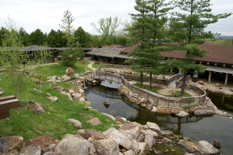 Arched wooden bridges connect a small island with tall pine trees in a stone rimmed pond to a grassy park area surrounded by low buildings