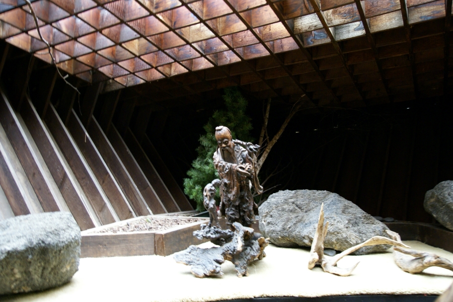 A statue carved out of driftwood depicts a human-like sea creature approaching a small malformed human. The piece is set in a sand bed surrounded by large rocks and branches below a skylight supported by wood framing