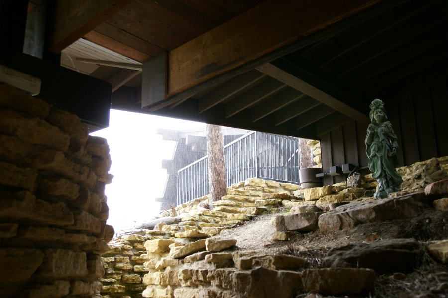 An aged bronze statue of a Chinese lady stands amongst stones below a wood framed roof while rain falls on a railed walkway and stones beyond