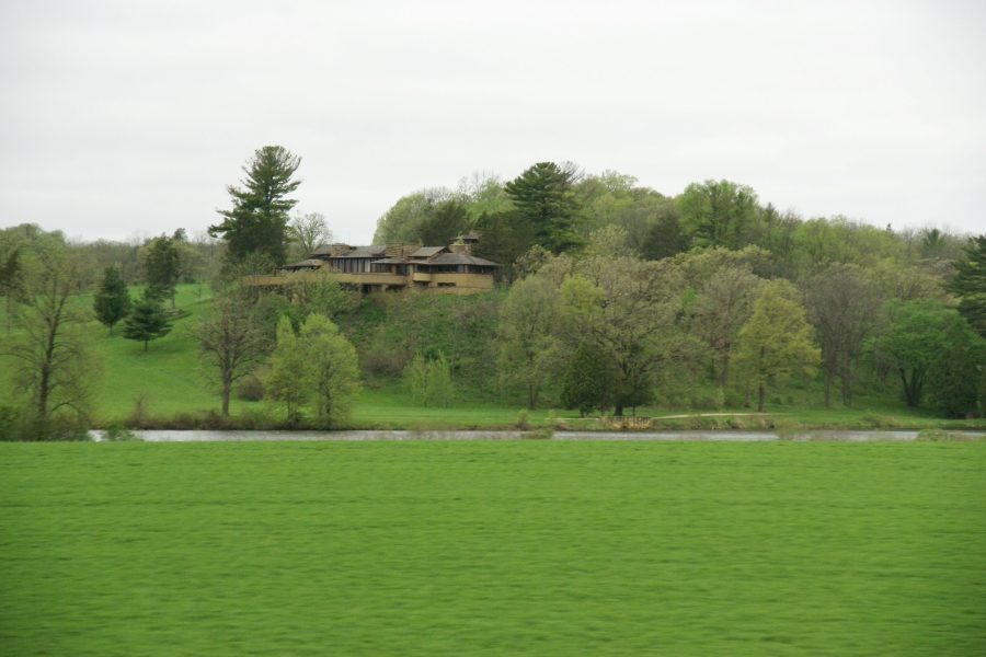 A house with yellow walls, large areas of glass and low sloped grayish brown roofs nestles into the trees on a hill above a stream