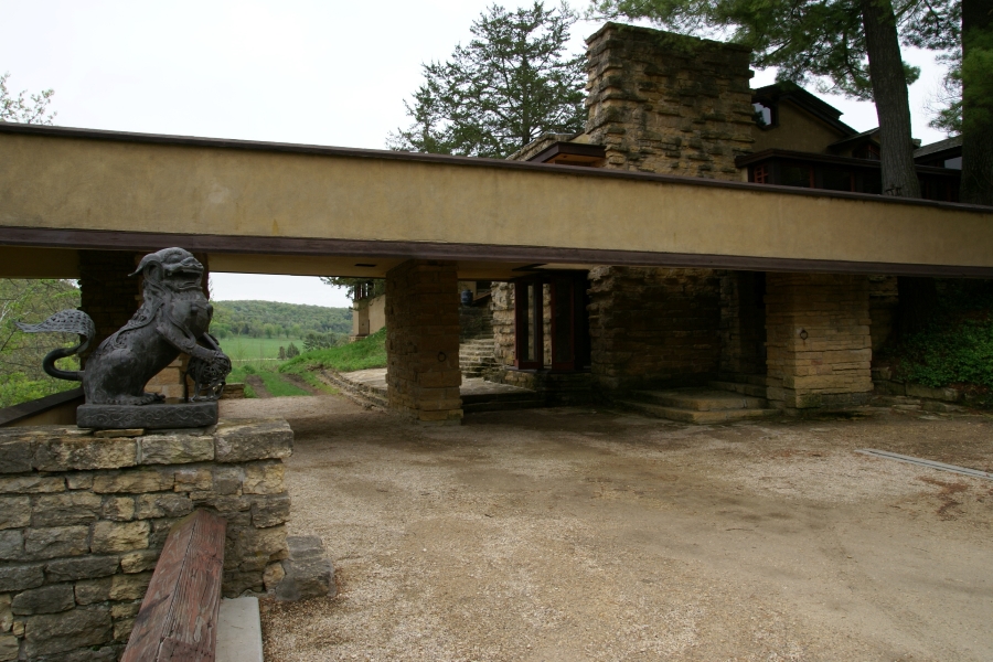 An oriental styled stone lion sits on a pedestal near a low covered area connecting to a large house